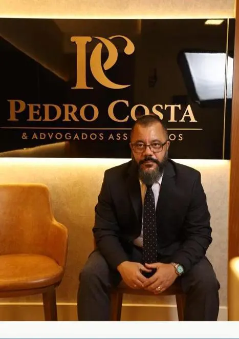 Dr. Pedro Costa sentado na recepção, diante da placa do escritório